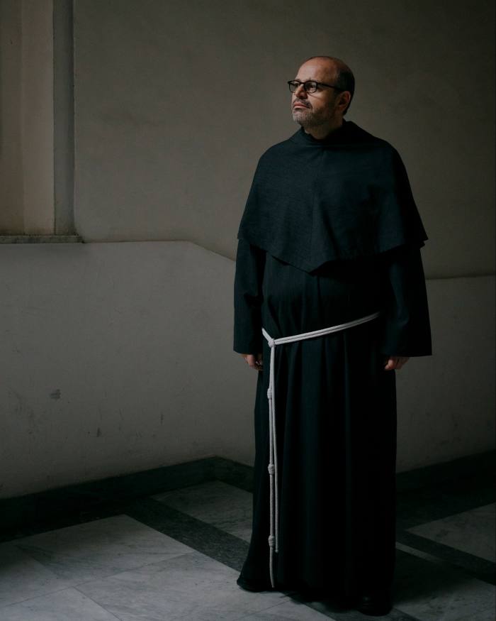 A full-length picture of Father Paolo Benanti standing in his habit against the plain walls of his monastery in Rome