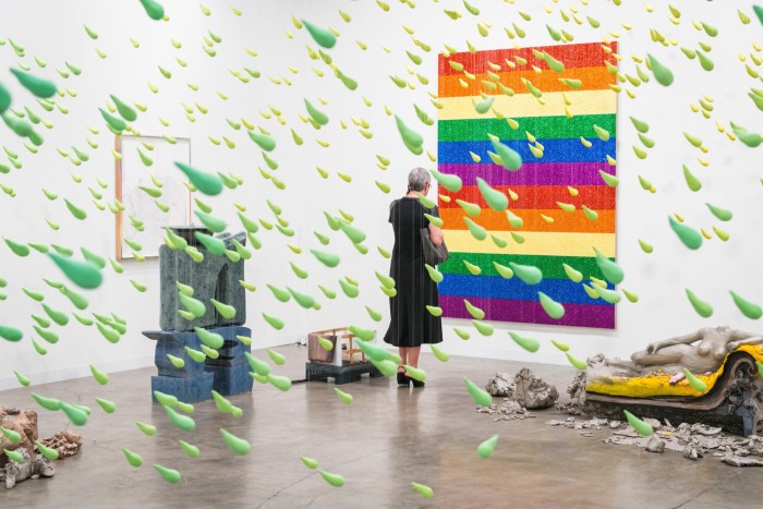 A woman looks at a rainbow-colored rectangular piece of art on a wall, while green plastic particles in the shape of raindrops shoot from the ceiling behind her