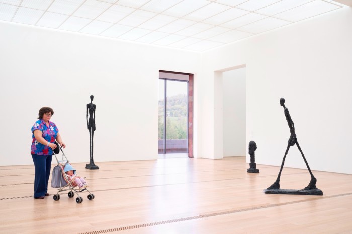 Two bronze stick figures and a bronze man's head sit in a large gallery while a woman with a pram watches from a distance