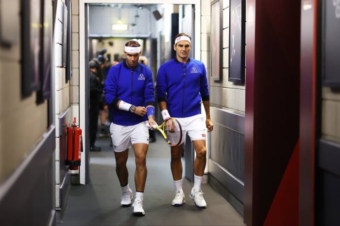 Nadal and Federer, in white shorts and blue zip-up tops, walk along a corridor
