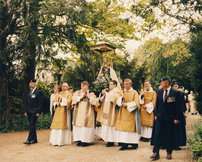 Priests in white and gold vestments walk along a treelined path, holding aloft the statue of Our Lady of Walsingham
