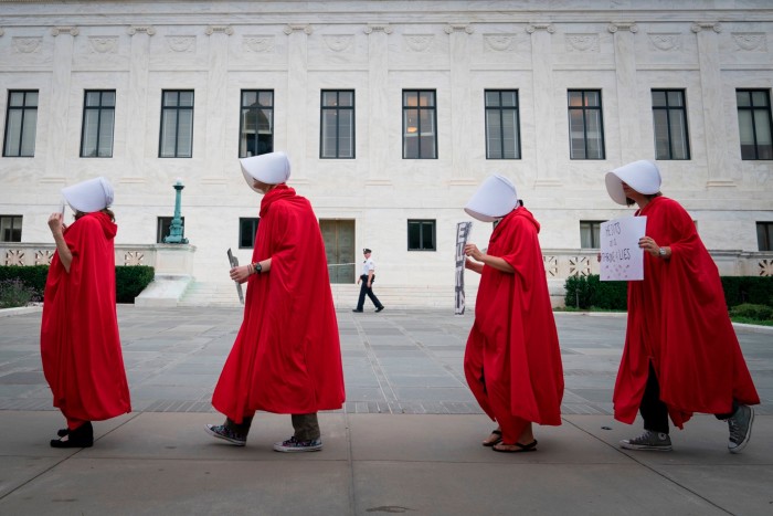 Four women dressed in red caps and white bonnets walk in a line, holding placards