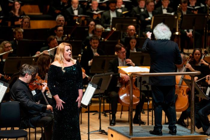 A white-haired man conducts an orchestra while a woman stands and sings