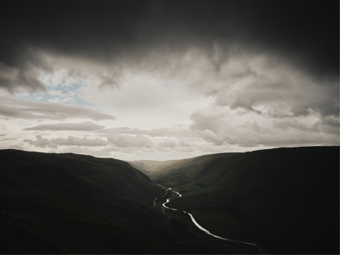 The sun glints through clouds over a landscape of hills with a road cutting through the valley