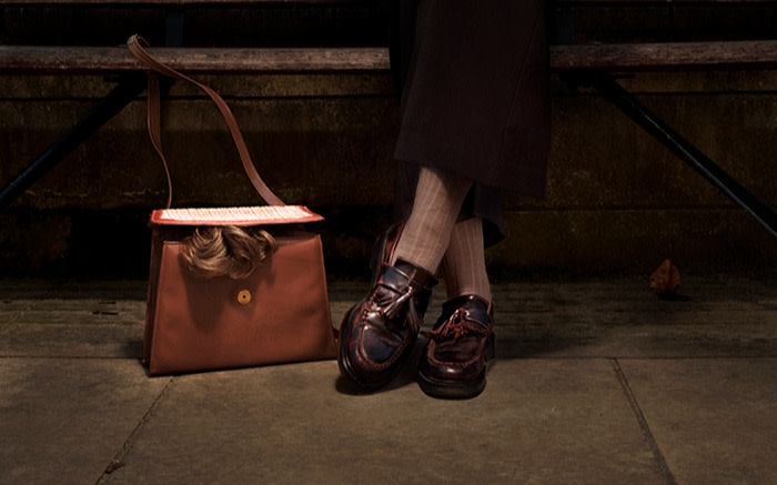 Shot of feet wearing loafers and a wig peeking out of a brown handbag 