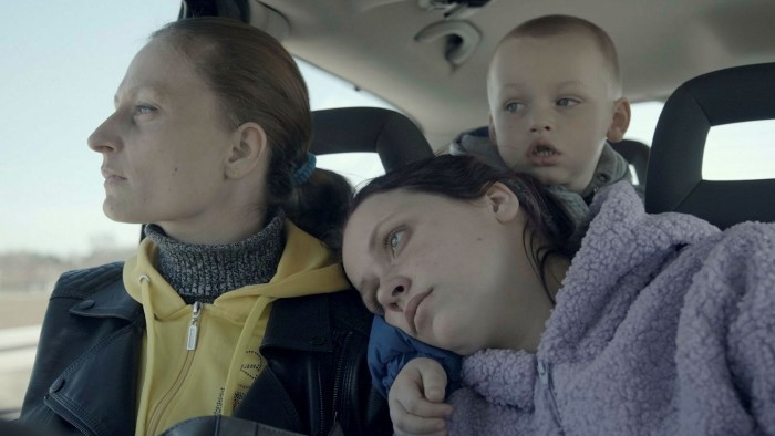 Moteris ir du vaikai sėdi automobilyje, važiuojančiame per kaimą, atrodo liūdni ir susirūpinę