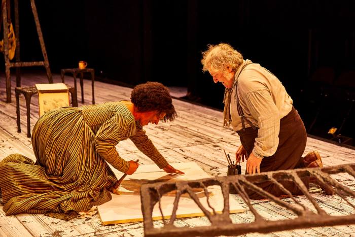 A woman and a man kneel on stage, working on a painting