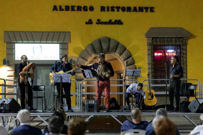 Several people in a row on a stage playing instruments in front of a yellow painted building