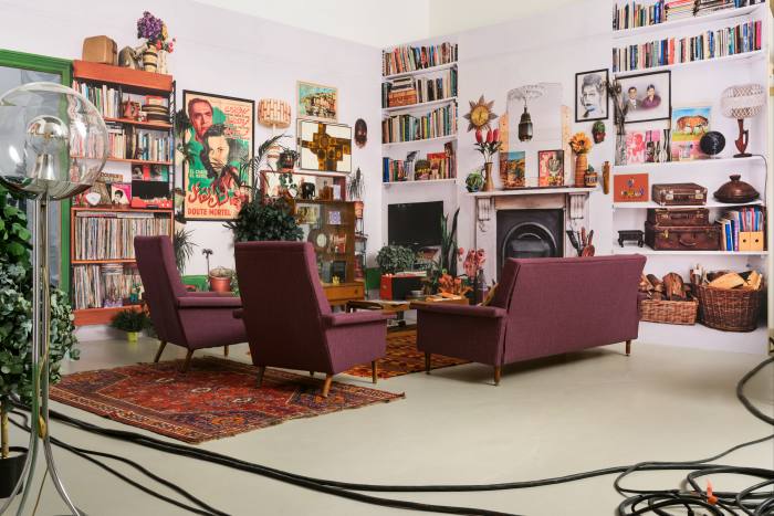 The interior of a living room, complete with purple sofa, bookshelves and posters for Arabic films, is recreated inside a white studio. Black cables run across the front of the picture.