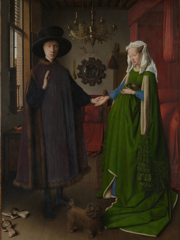 A portrait of a man and a woman standing holding hands. He holds up one hand, she rests a hand on her pregnant belly