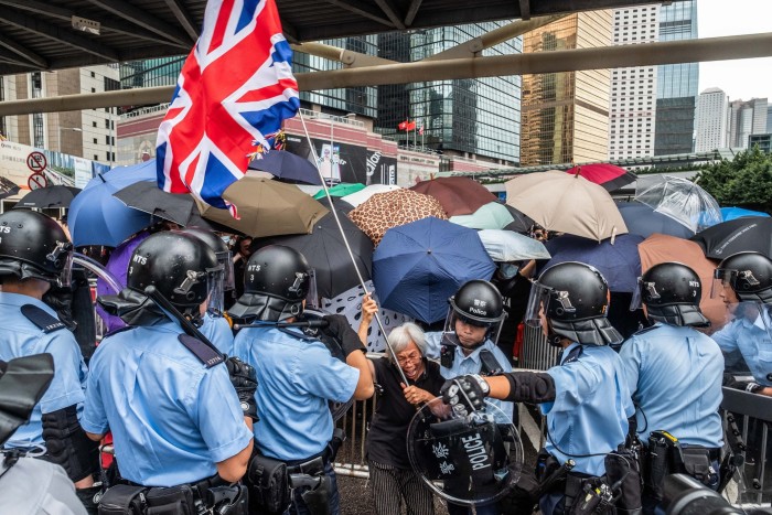 A woman holding waving a union jack is surrounded by police officers in riot gear, Behind her is a crowd of people carrying umbrellas