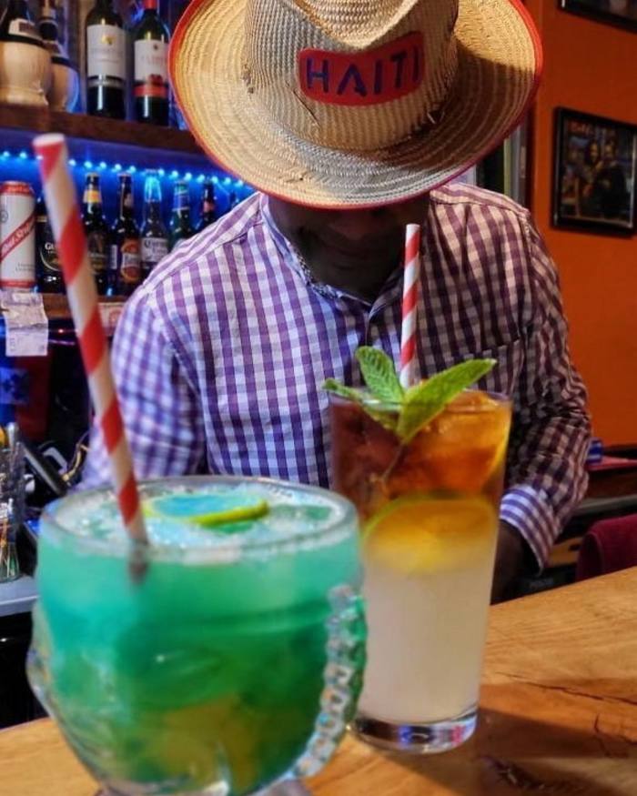 A man wearing a straw hat that says ‘Tahiti’ sitting behind two cocktails on at table at Grill Shack & Tiki Bar