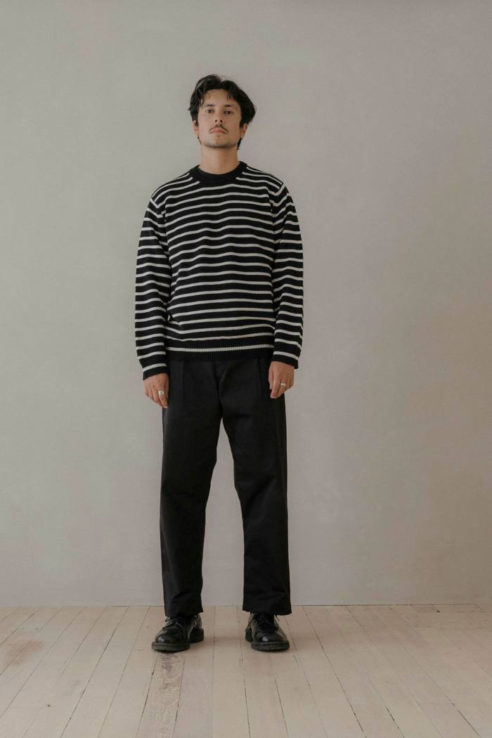 Evan Kinori stands against a white background. He has dark hair and wears a black and white striped top and dark trousers