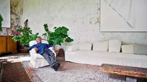 Axel Vervoordt in the Old Orangery, one of his favourite rooms in the  outer buildings of ‘s-Gravenwezel, his and his wife May’s 12th-century castle near Antwerp. Vervoordt designed the table from an old piece of wood. The painting above the sofa is by Japanese Gutai artist Norio Imai