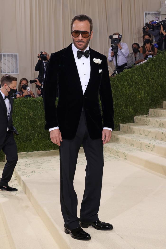 Tom Ford stands at the bottom of a flight of steps, with photographers behind him taking his picture.  He wears a suit with a bow tie and sunglasses