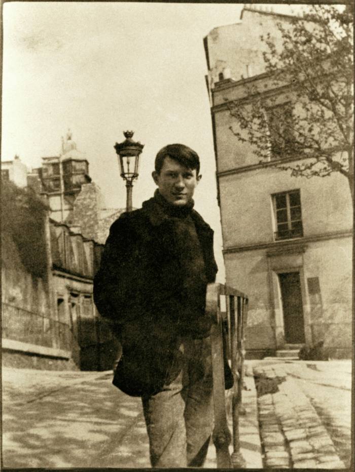 In a space in front of buildings, Picasso looks into the camera in dark casual clothes