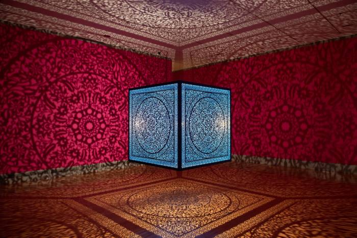 A bright blue glass box covered in an intricate pattern lit from within casts patterned shadows on the red walls