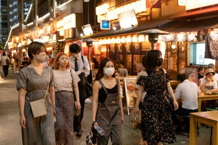 A group of people wearing face masks walk past restaurants at night in Shibuya, Tokyo