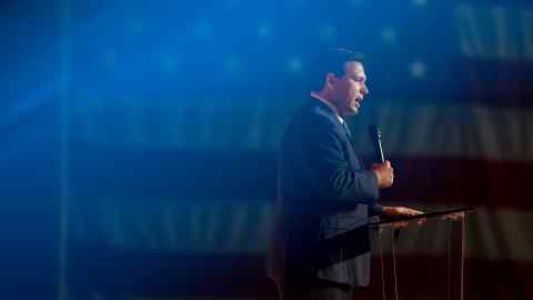 Ron DeSantis, gobernador de Florida, de pie en un podio con un micrófono en la mano.  Una bandera estadounidense gigante cuelga en el fondo