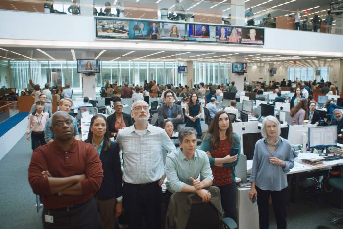 A group of journalists stare up at screens in a busy newsroom