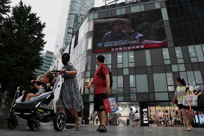 People walk in front of a large screen in Beijing showing a news broadcast about China’s military exercises encircling Taiwan