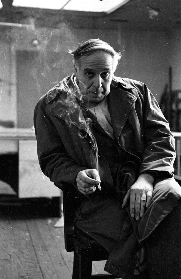 Guston is sitting in a chair, a cigarette in his hand.  He looks sullenly towards the camera, head down