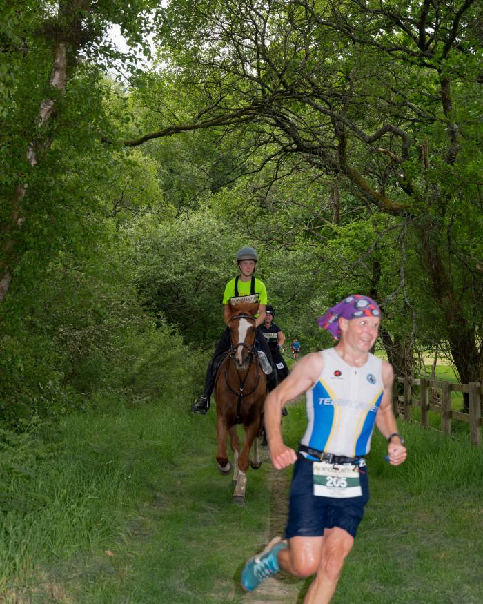 A runner in a bright headscarf runs through a wooded area with a horse close behind him