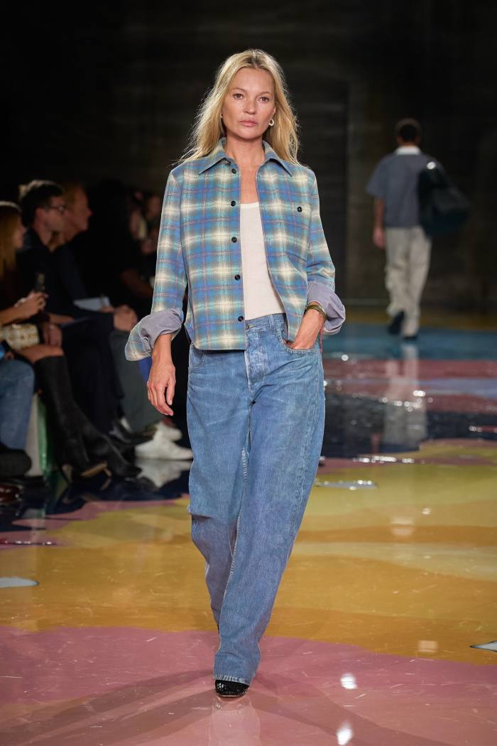 A blonde model on a catwalk wears what looks like baggy jeans and a plaid shirt over a white t-shirt