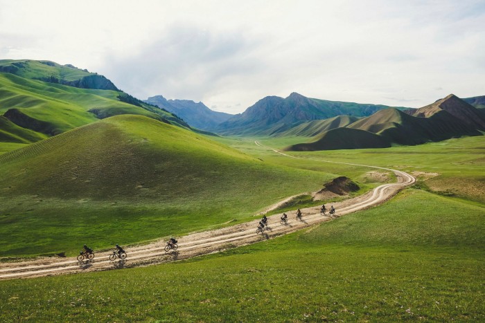 a line of cyclists ride on a dirt track across green hills, with mountains in the distance