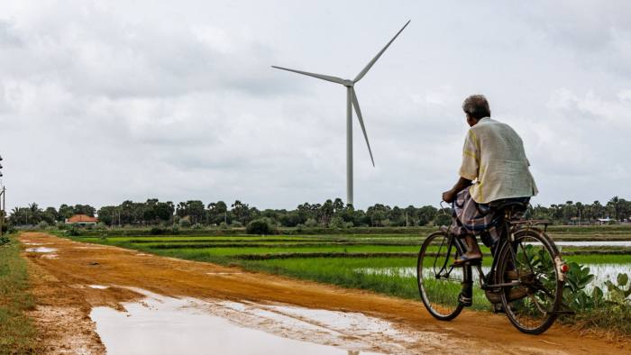 A man cycles away from us along a dirt road with a wind turbine looming in the distance