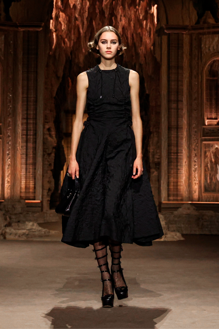 A model on a catwalk wears a black sleeveless below-the-knee dress