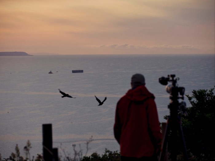 In the early morning light, a cameraman waits on the coast alongside his camera tripod 