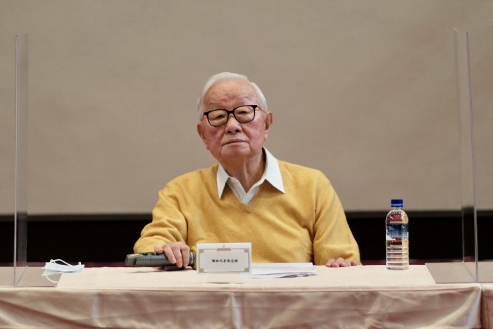 Morris Chang, wearing dark-rimmed glasses and yellow sweater with white collared shirt, sits at a table during a press conference