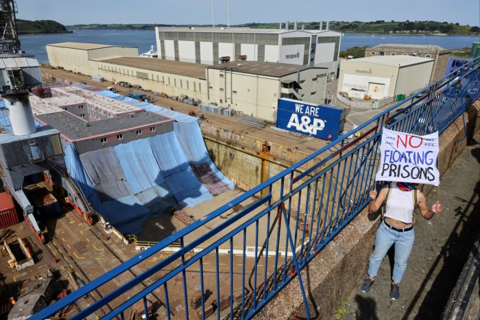 A woman stands on a walkway over a dry dock, holding up a sign that says No Floating Prisons