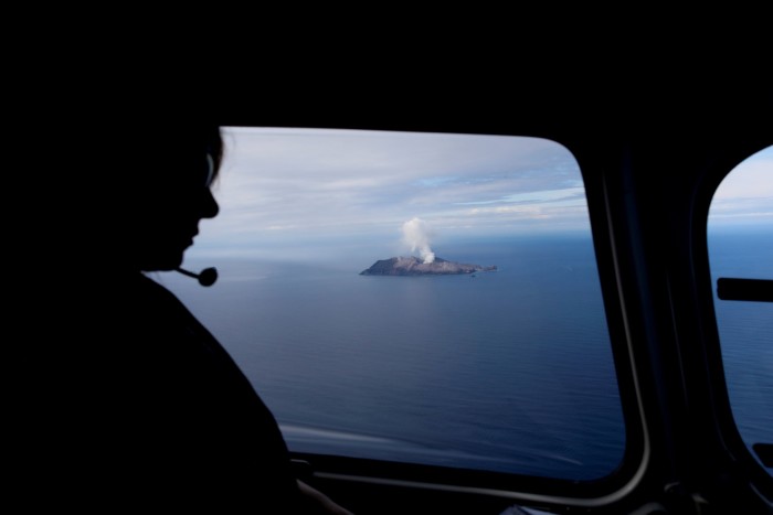 A woman looks out of an aircraft at smoke rising into the air from an island