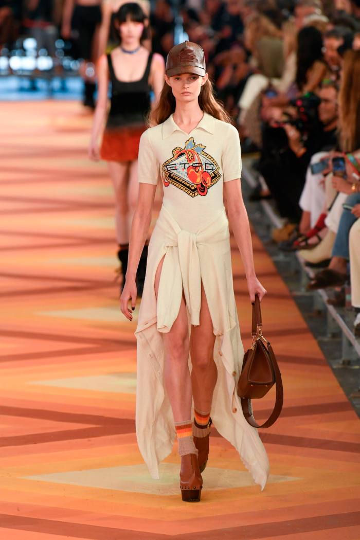 A model on a catwalk wears a dress with a hem that is short and fringed at the front, long at the back