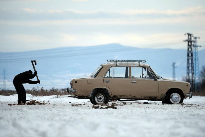 A boy chops wooden pallets in the snow next to a Lada car in Sofia, Bulgaria