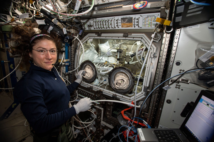 A woman astronaut wearing glasses and with her hair floating around her head operates a piece of equipment 