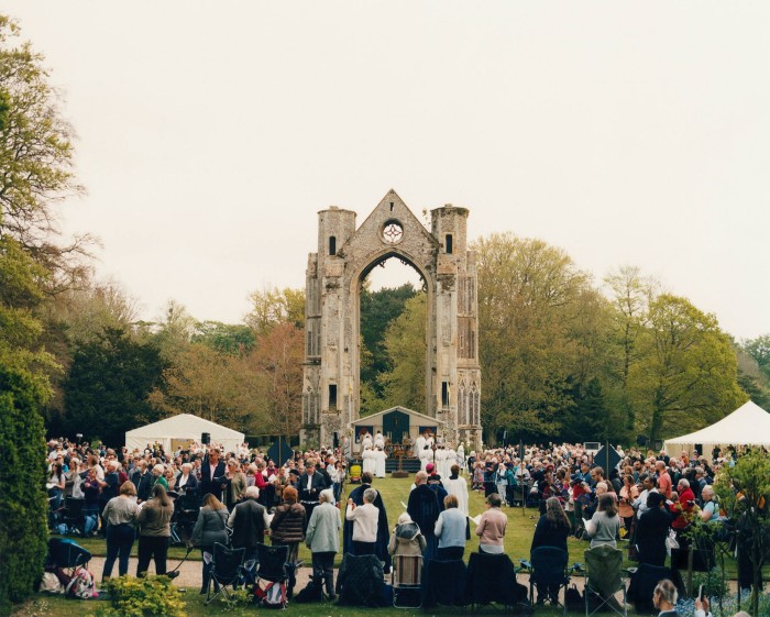Crowds of people stand near a stone arch, the remains of an old priory