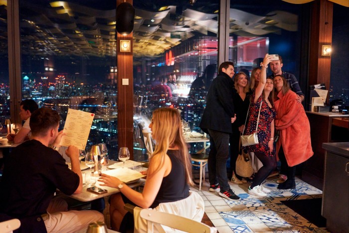 People stand near a window overlooking the city at night, smiling for the camera.  In front of them people dine at a table