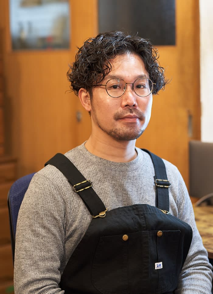 Ryoji Inoue, a bespectacled Japanese man with curly hair, mustache and beard, wearing a jumper outfit and undershirt