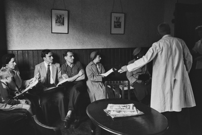 A black and white photograph of a doctor in a white coat gesturing to one of the people sitting in a waiting room
