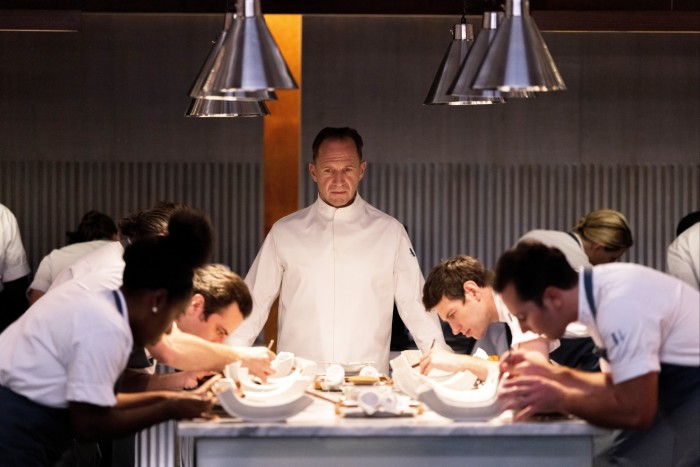A chef in white costume watches over a small army of cooks toiling over haute cuisine dishes