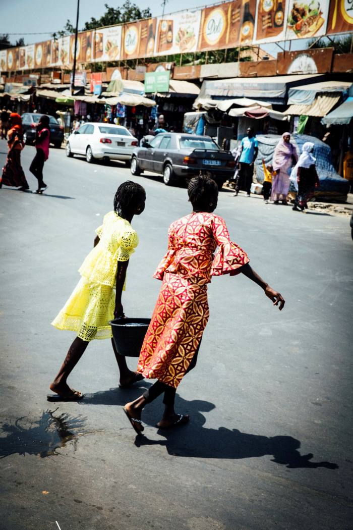 Two young girls, one in yellow, one in pink, carry a bucket of water between them as they cross a street in Dakar