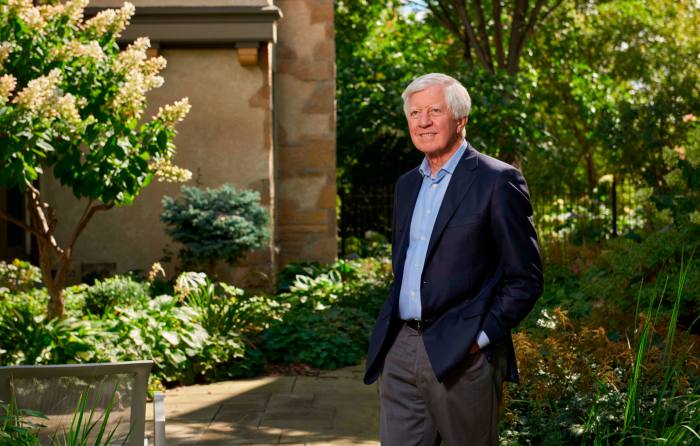 Bill George wearing a light blue collared shirt and navy blazer stands with his hands in his trouser pockets in a sunlit garden