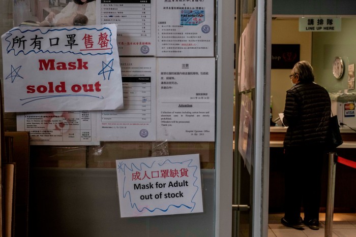 A woman in a mask stands at a reception desk in an office. On the wall outside, signs say ‘Mask sold out’