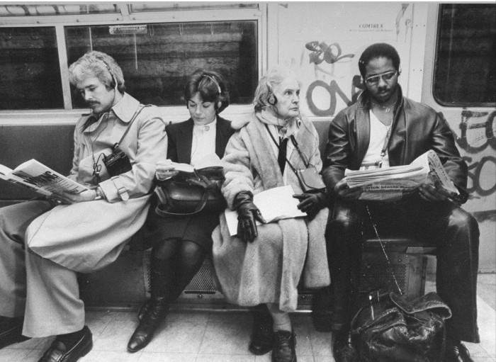 Black and white photo of two men and two women sitting in a New York subway car, three of them reading.  All wearable headphones for wearable listening devices