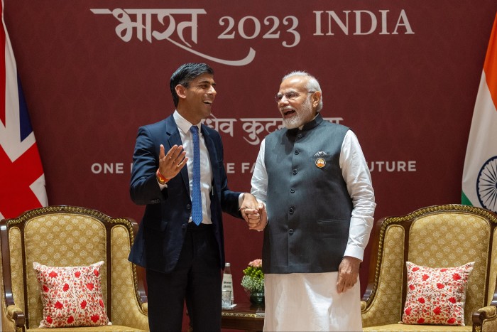 Rishi Sunak, in a suit and tie, alongside India’s prime minister Narendra Modi, who is wearing a long white robe and a green sleeveless jacket. The two are at the G20 summit in Delhi, India. Both men are smiling and clasping hands. National flags and two armchairs are on view in the background 