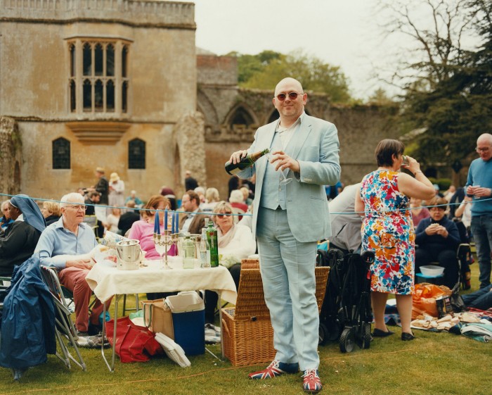 A man pours wine at an outdoor lunch near a church. behind him people eat and drink at picnic tables