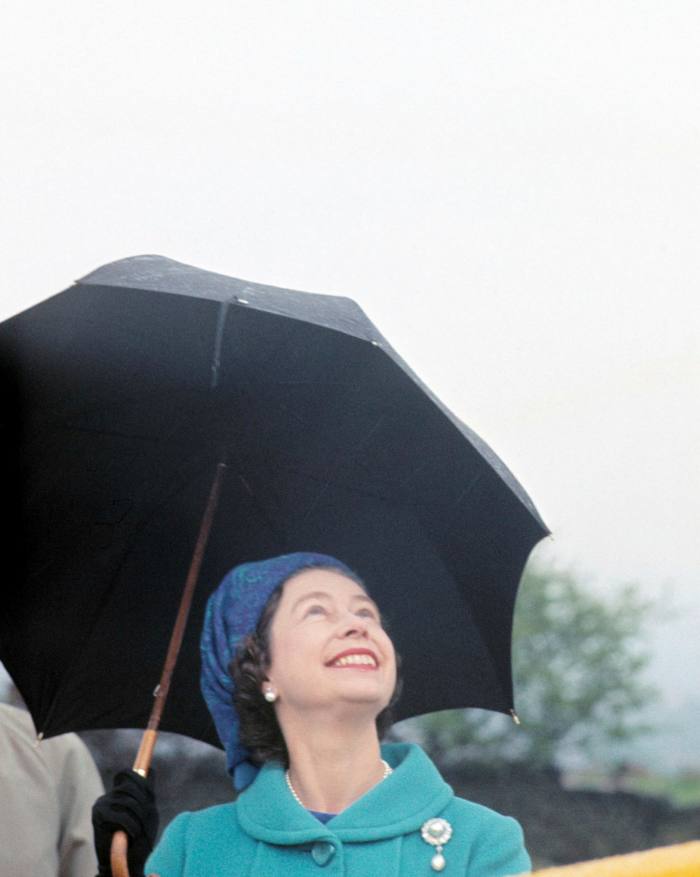 The queen holds a black umbrella and looks smiling towards the rainy sky